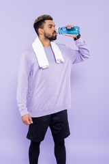 Wall Mural - bearded african american man in sweater standing with white towel and drinking water isolated on purple.