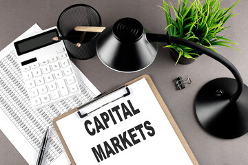 Poster - Clipboard ,chart and calculator, lamp and workspace accessories on a grey table. Top view , text CAPITAL MARKETS