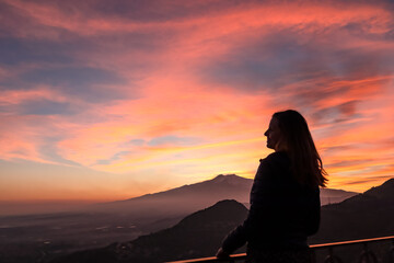 Wall Mural - Tourist woman watching beautiful sunset behind volcano Mount Etna near Castelmola, Taormina, Sicily, Italy, Europe, EU. Clouds with vibrant red orange colors. Silhouette of person during twilight