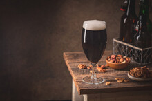 Glass Of Dark Beer With Foam Head On Dark Wooden Background With Empty Bottles And Beer Snacks