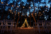 Festive String Lights Illumination On Boho Tipi Arch Decor On Outdoor Wedding Ceremony Venue In Pine Forest At Night. Vintage String Lights Bulb Garlands Shining Above Chairs At Summer Rural Wedding.