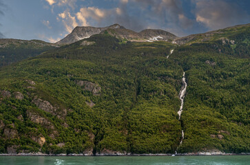 Wall Mural - Skagway, Alaska, USA - July 20, 2011: Taiya Inlet above Chilkoot Inlet. Wide landscape of forested mountain flank with white waterfall from source on top to green ocean water under blue cloudscape