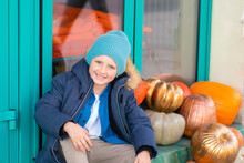 Boy In A Jacket Sitting On The Step  Near Pumpkins, Thanksgiving Decorations In Front Of The Entrance