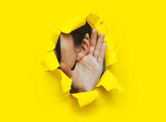 Wall Mural - Close-up of a left woman's ear and hand through a torn hole in the paper. Yellow background, copy space. The concept of eavesdropping, espionage, gossip and tabloids.