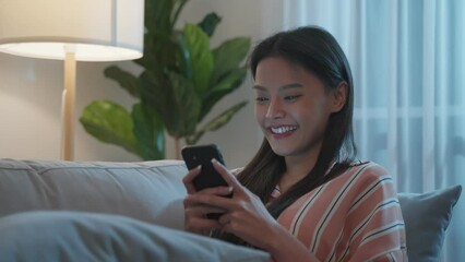 Wall Mural - Asian beautiful woman sit on sofa and chat on mobile phone at night.