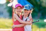 Fototapeta Łazienka - Two funny little girls of different races, in dresses and baseball caps, laughing and hugging. Best friends enjoy socializing and walking in the fresh air. Summer holidays