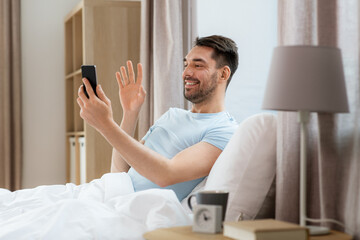 Wall Mural - technology, people and lifestyle concept - happy man with smartphone having video call in bed at home