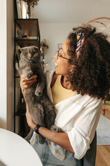 Cute young african girl holds fluffy gray cat in arms being distracted from work at laptop. Brunette wears glasses, shirt and admires her pet. Animal concept