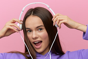 Wall Mural - a close horizontal portrait of a happy, enthusiastic young woman in purple clothes on a pink background wearing white wired headphones