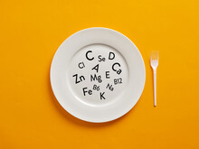 White Plate With Vitamin And Mineral Symbols And A Plastic Fork On Yellow Background. Healthy Nutrition, Immunity Maintenance And Diet
