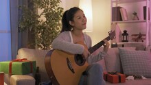 Asian Girl Celebrating Christmas Alone At Home Playing Guitar And Singing. Joyful Korean Girl Sitting With Gifts Around Is Holding Instrument And Watching Music Video Tape On Television