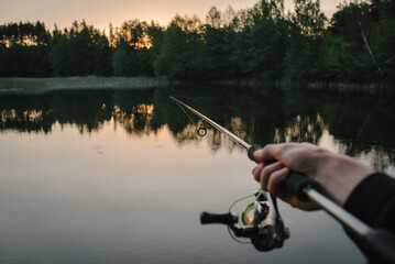 Wall Mural - Fisherman with rod, spinning reel on the river bank. Man catching fish, pulling rod while fishing from lake or pond with text space. Fishing for pike, perch on beach lake or pond. Fishing day concept.