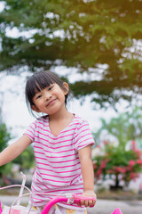 Wall Mural - Vertical image portrait of happy Asian child girl. She ride a bike at the green park garden. Cute girl with sweet smile. 4-5 years old childhood.