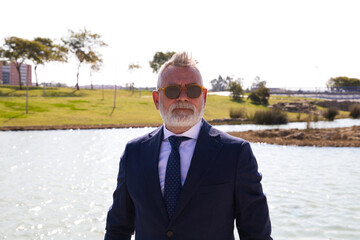 Handsome mature man, executive, grey hair, beard, sunglasses, jacket and tie, is in a park with an artificial lake posing for photo. Concept of mature man, grandfather, executive.