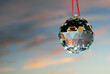 A faceted crystal ball hanging on a red string reflecting the light and colours of the evening sky with pink clouyds on the background. Horizontal