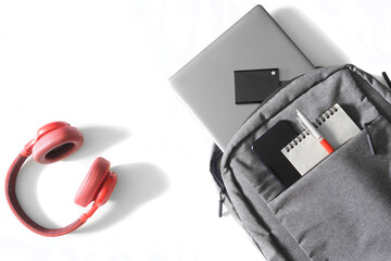 Gray urban backpack or bag with laptop, mobile phone, external ssd disk, notepad and pen lies on a white table next to red headphones and mouse. Tools and gadgets of a student or freelancer.