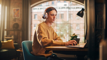 Poster - Young Handsome Man Working from Home on Desktop Computer in Sunny Stylish Loft Apartment. Creative Designer Wearing Cozy Yellow Sweater and Headphones. Urban City View from Big Window.