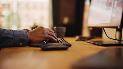 Poster - Close Up Footage of Person's Black Hands Use Computer while Sitting at a Table. Creative Designer in a Grey Shirt Using Mouse, Typing on Keyboard. Monitor Display and Laptop on a Desk.