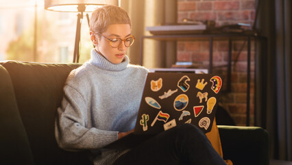 Wall Mural - Stylish Female Using Laptop Computer with Diverse LGBT and Lifestyle Stickers on the Back. Young Creative Woman Sitting on a Couch, Typing, Browsing Internet and Checking Social Media at Home.