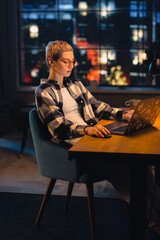 Fototapete - Vertical Screen: Young Attractive Woman Using Laptop Computer in Stylish Loft Apartment in the Evening. Creative Female Checking Social Media, Browsing Internet, Wearing Cozy Clothes.