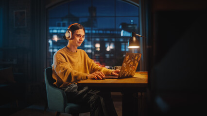 Wall Mural - Young Handsome Man Opening Laptop Computer in Stylish Loft Apartment in the Evening. Creative Person Wearing Cozy Yellow Sweater and Putting On Headphones. Urban City View from Big Window.