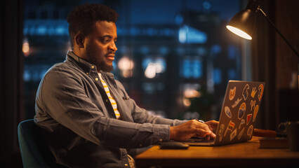 Poster - Young Handsome Black Man Working from Home on Laptop Computer in Stylish Loft Apartment in the Evening. Creative Male Gets a Great Idea while Checking Social Media. Urban City View from Big Window.
