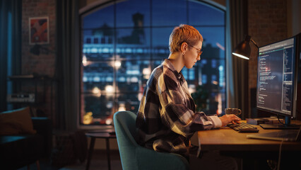 Fototapete - Young Woman Writing Code on Desktop Computer in Stylish Loft Apartment in the Evening. Creative Female Wearing Cozy Clothes, Working from Home on Software Development. Urban City View from Big Window.