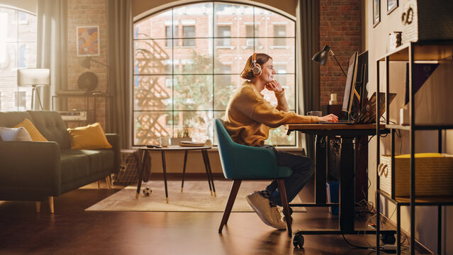 Fototapete - Young Handsome Man Working from Home on Desktop Computer in Sunny Stylish Loft Apartment. Creative Designer Wearing Cozy Yellow Sweater and Headphones. Urban City View from Big Window.