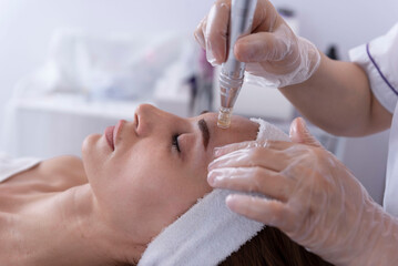 close up of Cosmetologist,beautician applying facial dermapen treatment on face of young woman customer in beauty salon.Cosmetology and professional skin care, face rejuvenation.