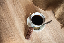 A Cup Of Fragrant Black Coffee On A Saucer With A Spoon And A Chocolate Bar Lie On A Wooden Brown Table Covered With Linen.