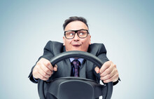 Portrait Of A Comical Businessman In A Dark Suit And Glasses Holding A Car Steering Wheel In His Hands, On A Blue Background