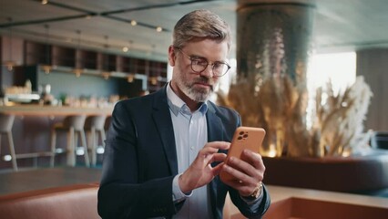 Shot of handsome smiling man in formal outfit using smartphone in restaurant, touching screen of mobile phone, scrolling social media. Communication. Indoors. Business people. Working day. Summertime