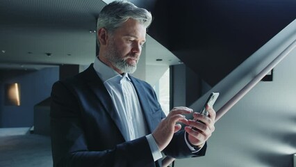 Footage of confident middle-aged Caucasian businessman networking on cellphone, standing near stairs, slow motion. Shot of serious good-looking gray-haired man in suit holding smartphone in hands