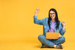 Leinwandbild Motiv Happy winner! Business concept. Portrait of happy young woman in casual sitting on floor in lotus pose and holding laptop isolated over yellow background.