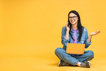 Business concept. Portrait of happy young woman in casual sitting on floor in lotus pose and holding laptop isolated over yellow background. Using mobile phone.