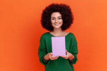 Wall Mural - Portrait of satisfied woman with Afro hairstyle in green casual style sweater standing showing paper notebook, looking at camera, happy expression. Indoor studio shot isolated on orange background.