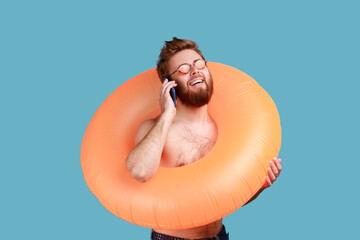 Portrait of overjoyed young adult bearded man standing with orange rubber ring and talking phone, telling about his excellent vacation. Indoor studio shot isolated on blue background.