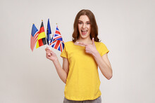 Positive Smiling Teenager Girl In Yellow Casual T-shirt Holding Flags Of Usa, German, Great Britain And Europe Countries, Showing Thumb Up. Indoor Studio Shot Isolated On Gray Background.