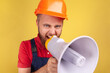 © khosrork - Angry aggressive bearded worker wearing protective helmet and blue overalls holding megaphone and screaming, point of view photo. Indoor studio shot isolated on yellow background.