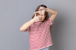 Portrait of little girl wearing striped T-shirt looking through fingers imagining frame to make good photos, dreaming to be a photographer. Indoor studio shot isolated on gray background.