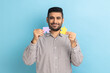 Portrait of optimistic bearded businessman holding yellow and purple puzzle pieces, solving tasks, looking at camera, wearing striped shirt. Indoor studio shot isolated on blue background.