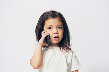 Wall Mural - a little girl of preschool age, sitting on a white background in a white T-shirt, enthusiastically talking on the phone, turning half-side to the camera. The topic of the conversation on the phone