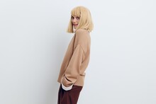  Horizontal Photo, A Woman On A White Background In A Beige Sweater And Brown Trousers With Fine Blonde Hair Stands Sideways And Smiles With Her Hands In Her Pockets