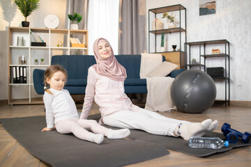 Wall Mural - Young smiling muslim mother doing sports exercise at home, wearing hijab, together with funny little daughter, enjoying activities with baby, fun and sport practice. Healthy lifestyle concept photo.