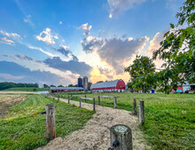 A Gravel Cow Lane With A Dairy Barn And Two Silos And Storm Clouds At Sunset In The Summer.