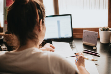 Close up detail image of a young woman studying and working on his home desk, doing homework during university, preparing for exam with textbook and taking notes, selective focus on the pencil.