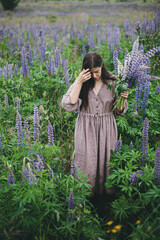 Wall Mural - Stylish woman in rustic dress holding lupine bouquet in meadow. Cottagecore aesthetics. Young female in linen dress gathering wildflowers in atmospheric summer countryside, rural slow life