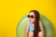 Summer vacation by the sea. A cute girl in sunglasses and a swimsuit with an inflatable swimming circle on a yellow background. Space for text