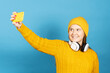 © Egoitz - Cute blonde teenager girl taking selfie isolated on blue background. Young woman wearing yellow winter hat and jersey