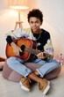 © Seventyfour - Vertical full length portrait of black teenage boy playing guitar and smiling at camera while sitting on bean bag